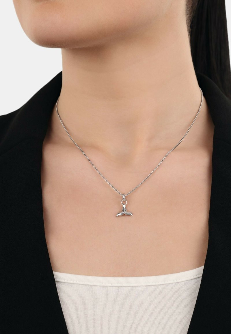 Pendente a forma di coda di balena in argento su una catena sottile. Presenta una superficie lucida e un design delicato. Indossato sopra un top di colore chiaro.