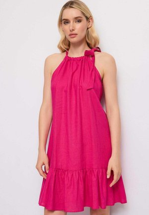 Donna bionda che indossa un abito senza maniche rosa acceso con collo all'americana e fiocco annodato vicino allo scollo, in piedi davanti a uno sfondo bianco semplice.