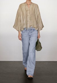 Blouse oversize beige clair texturée avec un col rond et des liens à l'avant, portée avec un jean bleu clair à jambes larges et un sac à main vert.