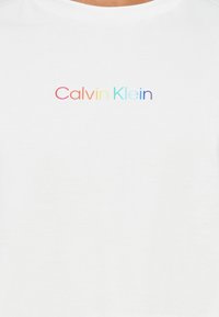 Valkoinen puuvillapaita, jossa on keskellä rinnassa "Calvin Klein" -logo, joka on gradienttiväreissä (punaisesta purppuraan). Lyhyet hihat ja pyöreä kaula-aukko.
