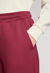 Burgundske atletske trenirke s mekanom teksturom, elastičnim pojasom i bočnom džepom, u kombinaciji s dugim kremastim topom.
