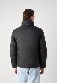 Svart polstret jakke med høy krage og glidelås. Har en glatt, blank overflate og er paret med lyseblå denimjeans.