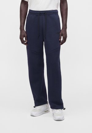 Homme portant un pantalon de jogging bleu marine à cordon et des baskets blanches, debout devant un fond clair uni.