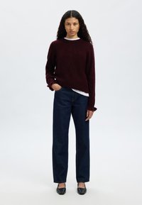 Burgundski pleteni pulover z dolgimi rokavi, nošen čez belo srajco, kombiniran s temno modrimi kavbojkami z ravnimi hlačnicami in črnimi balerinkami z ostrim sprednjim delom.