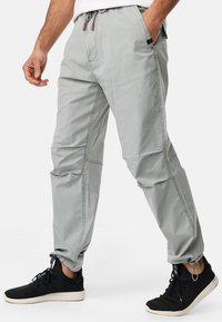Pantalon gris clair en coton avec une taille élastique, un cordon de serrage, des poches latérales et des jambes fuselées. Associé à des chaussures de sport noires.