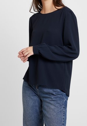 Vrouw die een marineblauwe blouse met lange mouwen en gerimpelde manchetten draagt, gecombineerd met een blauwe spijkerbroek, staand met zachtjes gevouwen handen.