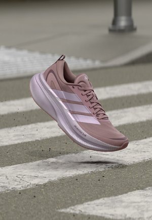 Zapatilla de correr rosa de Adidas con rayas blancas suspendida sobre un paso de peatones en una calle asfaltada gris.