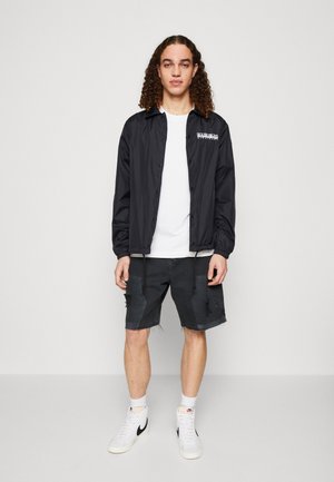 Ung mand med krøllet hår iført en sort knapjakke, hvid t-shirt, slidte sorte shorts, hvide sokker og hvide Nike-sneakers.