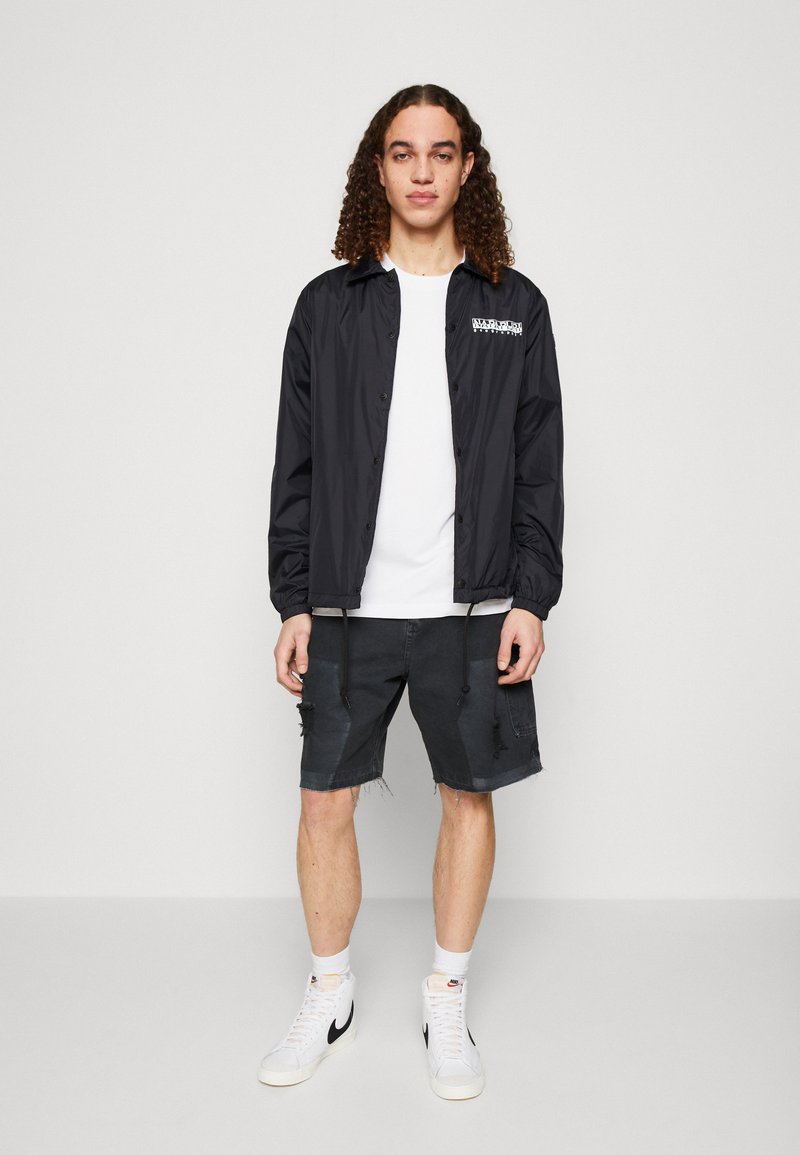 Giovane con i capelli ricci che indossa un giubbotto nero con bottoni, una maglietta bianca, pantaloni corti neri strappati, calzini bianchi e sneakers bianche Nike.