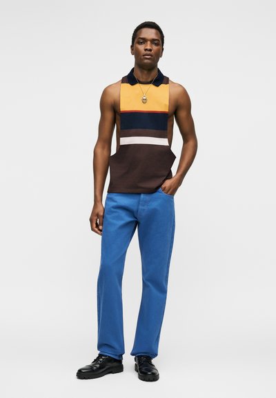 Νέος άνδρας που φοράει αμάνικο πόλο με χρωματικούς συνδυασμούς, μπλε τζιν, μαύρα παπούτσια και μενταγιόν, στέκεται με το ένα χέρι στην τσέπη.