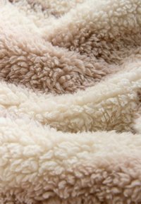 Miękki, teksturowany pled w odcieniach beżu i kremu, z puszystą, włochatą powierzchnią o falistym, warstwowym wyglądzie.