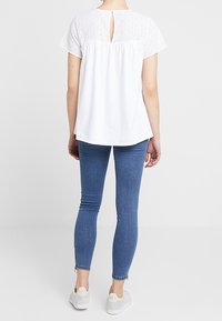 Biała bluzka z detalem z ażurowej koronki na rękawach, z odsłoniętymi plecami z wycięciem typu keyhole, zestawiona z niebieskimi obcisłymi dżinsami i jasnymi tenisówkami.