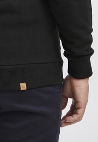 Sudadera negra con puños y dobladillo acanalados. Presenta una tela texturizada y una pequeña etiqueta de cuero marrón con un logo cerca del dobladillo.