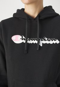 Čierna mikina s kapucňou s farebným logom "Champion" v ružovej a bielej farbe, s nastaviteľnými šnúrkami a prednou kapsou. Mäkka textúra.
