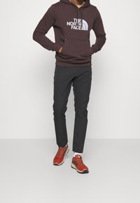 Felpa marrone scuro con logo bianco, pantaloni neri e scarpe da ginnastica arancio-rosse. La felpa presenta una tasca a marsupio e un cappuccio regolabile.