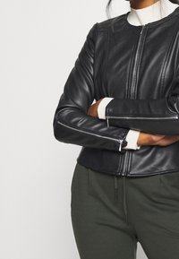 Persona che indossa una giacca di pelle nera con zip sopra un maglione bianco, con le braccia incrociate e pantaloni verde scuro con coulisse.