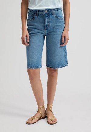 Kvinde iført blå jeansshorts i mellemlang længde, hvid top og gyldne flade sandaler med stropper, stående mod en ensfarvet baggrund.