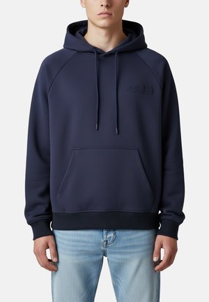 Uomo che indossa una felpa con cappuccio blu navy con tasca frontale e coulisse, abbinata a jeans azzurri chiari, in piedi davanti a uno sfondo semplice.