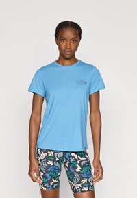 Μπλε μπλουζάκι με στρογγυλή λαιμόκοψη και κοντά μανίκια, με μικρό τυπωμένο λογότυπο στο αριστερό στήθος. Συνδυάζεται με σορτς σε μαύρο χρώμα με πολύχρωμο μοτίβο.