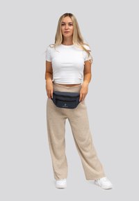 Marsupio nero con due scomparti con zip, indossato in vita sopra pantaloni a gamba larga beige chiaro. Tessuto testurizzato con dettaglio del logo.