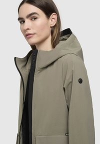 Jachetă ușoară, de culoare khaki, cu glugă, fermoar negru, buzunare frontale mari și detaliu circular cu logo pe mânecă. Textură netedă.