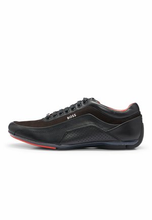 Zwarte lage leren sneaker met geperforeerd zijpaneel, vetersluiting aan de voorkant, rode accenten op de zool en "BOSS"-branding aan de zijkant.