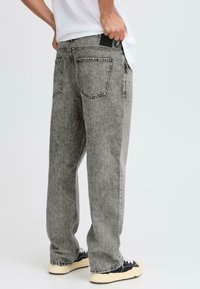 Persona vestida con unos vaqueros de acid-wash gris de corte holgado, una camiseta blanca y zapatillas negras con suelas beige, de pie frente a un fondo sencillo.