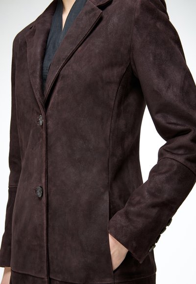 Personne portant un blazer en daim marron foncé avec des boutons, une main dans la poche, montrant un gros plan du haut du torse.