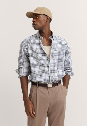 Checked button-up shirt in light blue, white, and brown; rolled-up sleeves; paired with beige cap and brown belt, cream tank top underneath.