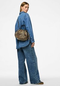 Kabelka s leopardím vzorem, se stahovací šňůrkou, texturovaným materiálem a dvojitými rukojeťmi. Modelka má na sobě modrou džínovou košili a široké kalhoty.