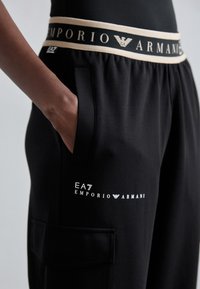 Melni sporta bikses ar platu beigu krēmkrāsas jostu, uz kuras melnā krāsā ir rakstīts "EMPORIO ARMANI", kā arī logo uz apakšējās kājas. Iekļautas sānu kabatas.