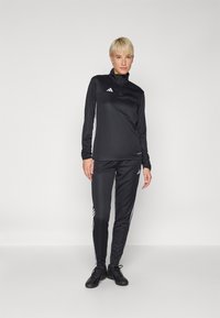 Schwarzer Sportanzug mit Reißverschlusskragen, mit weißen Streifen an den Ärmeln und den Seiten. Hergestellt aus glattem Stoff. Schwarze Sportschuhe, Stehpose.