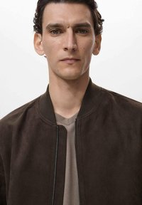 Dunkelbraune Wildleder-Bomberjacke mit geripptem Kragen und Reißverschluss, getragen über einem hellbeigen V-Ausschnitt-Shirt. Minimalistisches Design.