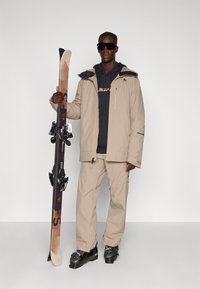 Beige skijack en bijpassende broek, zwarte hoodie. Houdt ski's met zwarte bindingen vast. Donkere zonnebril maakt de look compleet.
