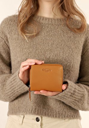 Tan leather wallet with smooth texture, zip closure, and a logo embossed on the front, held in hands, with a knitted beige sweater in background.