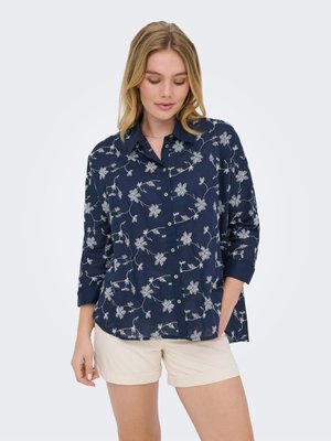 Femme portant une chemise bleu marine à boutons avec un motif floral blanc et un short beige, debout avec une main dans la poche, regardant vers le bas.