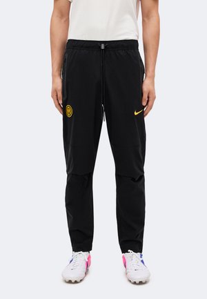 Άτομο που φοράει μαύρη φόρμα Nike με κίτρινα λογότυπα και λευκά ποδοσφαιρικά παπούτσια με ροζ και μπλε λεπτομέρειες σε λευκό φόντο.