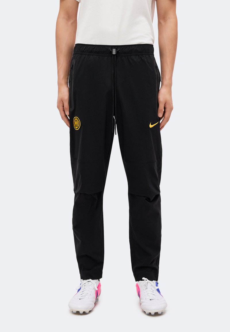 Άτομο που φοράει μαύρη φόρμα Nike με κίτρινα λογότυπα και λευκά ποδοσφαιρικά παπούτσια με ροζ και μπλε λεπτομέρειες σε λευκό φόντο.