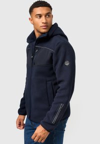Tumed fleece'i jakk avatud ees, kapuutsi ja kahe eesmine taskuga. Omab tekstuurset keha ja siledat paneeli rinnal ning varrukatel.