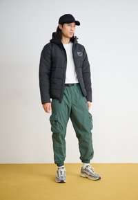 Μαύρο επενδυμένο μπουφάν με κουκούλα και λογότυπο, λευκό μπλουζάκι, πράσινα cargo παντελόνια και γκρι αθλητικά παπούτσια με έντονες λεπτομέρειες. Στέκεται σε κίτρινο δάπεδο.