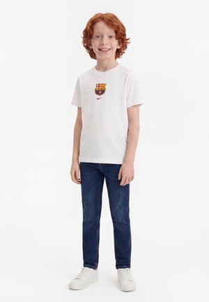 Rødhåret dreng smiler, iført en hvid t-shirt med et farverigt våbenskjold, blå jeans og hvide sneakers, stående mod en ensfarvet hvid baggrund.