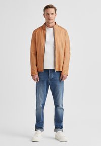 Sandyfarbene Lederjacke mit Stehkragen, kombiniert mit einem weißen T-Shirt und blauen Jeans. Weiße Sneaker runden das Outfit ab. Schlichtes Design.