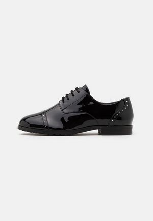 Chaussure de ville pour homme en cuir verni noir avec bout cap, lacets, petites rivets argentés et talon bas sur fond blanc.