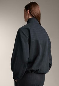 Fekete tűcsíkos zakó magas gallérral, bő szabással és rugalmas mandzsettákkal. Puha textúra finom, kontrasztos csíkmintákkal.