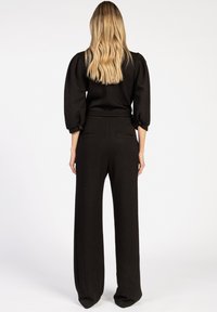 Zwarte jumpsuit met pofmouwen, getailleerde bodice en wijde broekspijpen. Zacht materiaal met subtiele textuur; beschikt over achterzakken en een riem.