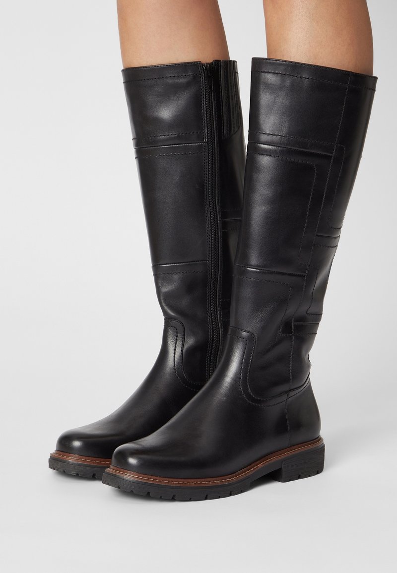 Paire de bottes en cuir noires jusqu'aux genoux avec fermeture éclair latérale, surpiqûres marron sur la semelle, et semelles en caoutchouc épaisses portées par une personne.