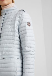 Svetle modrá prošívaná bunda s vysokým límcem a logem na levém rukávu. Obsahuje boční kapsu se zipem na uchování věcí.