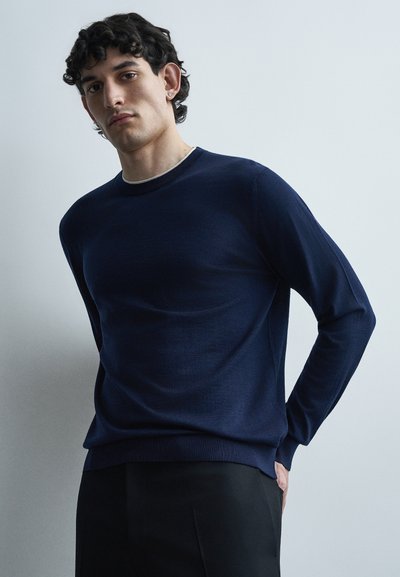 Jeune homme aux cheveux bouclés et foncés portant un pull bleu marine et un pantalon noir, debout avec les mains derrière le dos sur un fond uni.