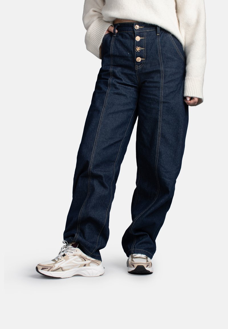 Persona che indossa jeans a vita alta a gamba larga di colore blu scuro con quattro bottoni, maglione bianco e sneaker bianche e beige, in piedi con una mano in tasca.