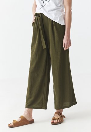 Persoon draagt olijfgroene wijd uitlopende broek met een gestrikte tailleband, wit overhemd en bruine open teen sandalen, staand op een witte vloer.